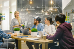 A group of 5 adults brainstroming and sharing an ideas around a table and writing them on a big chalkboard