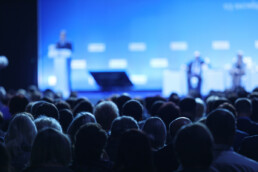 Focused image on a large crowds heads whop are at a conference with someone speaking on a podium